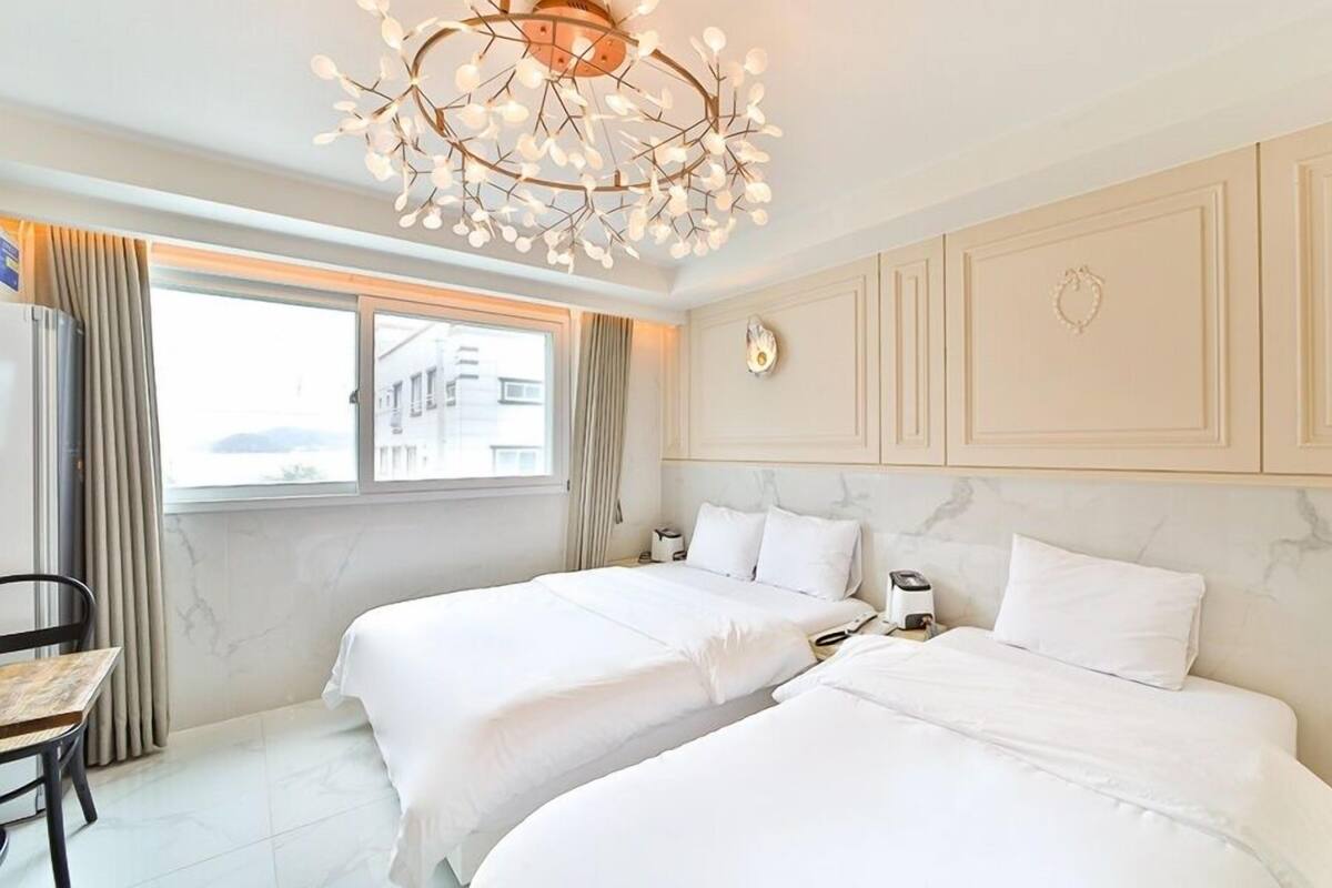 A hotel room with two beds, a chandelier, a desk, and a window with curtains.