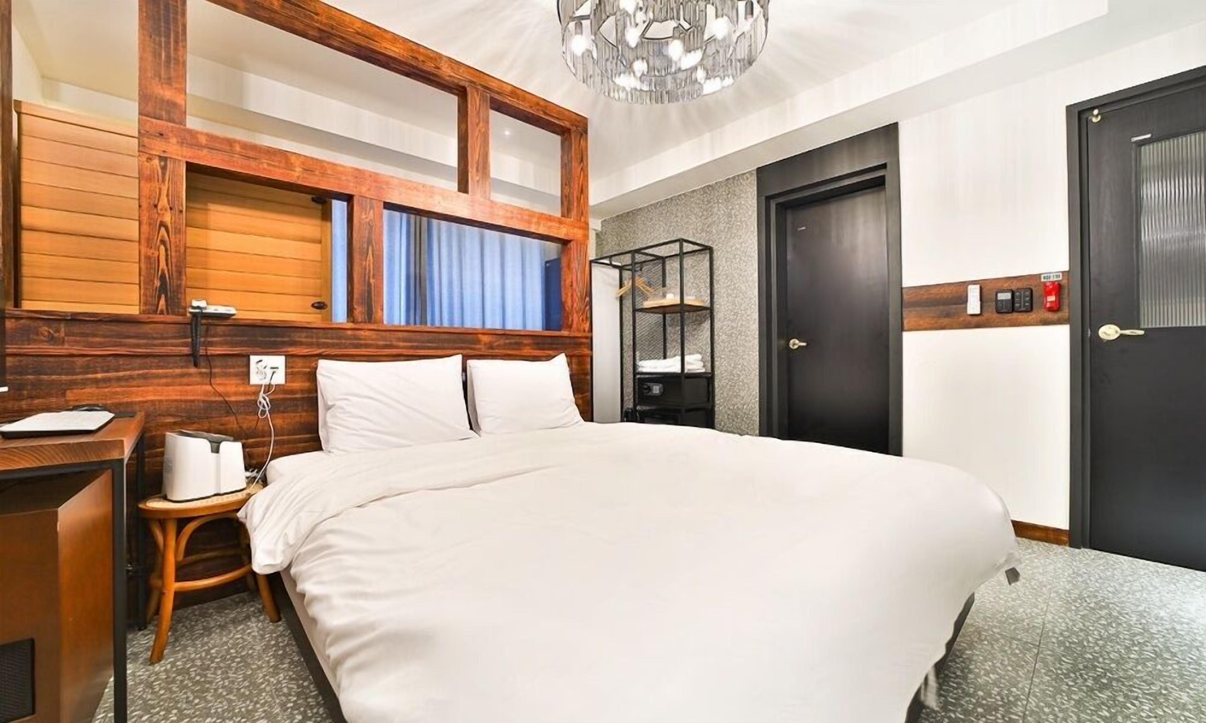 A hotel room with a large bed, wooden headboard, a desk with a chair, a small side table, a wardrobe, and a door with a key in the lock.
