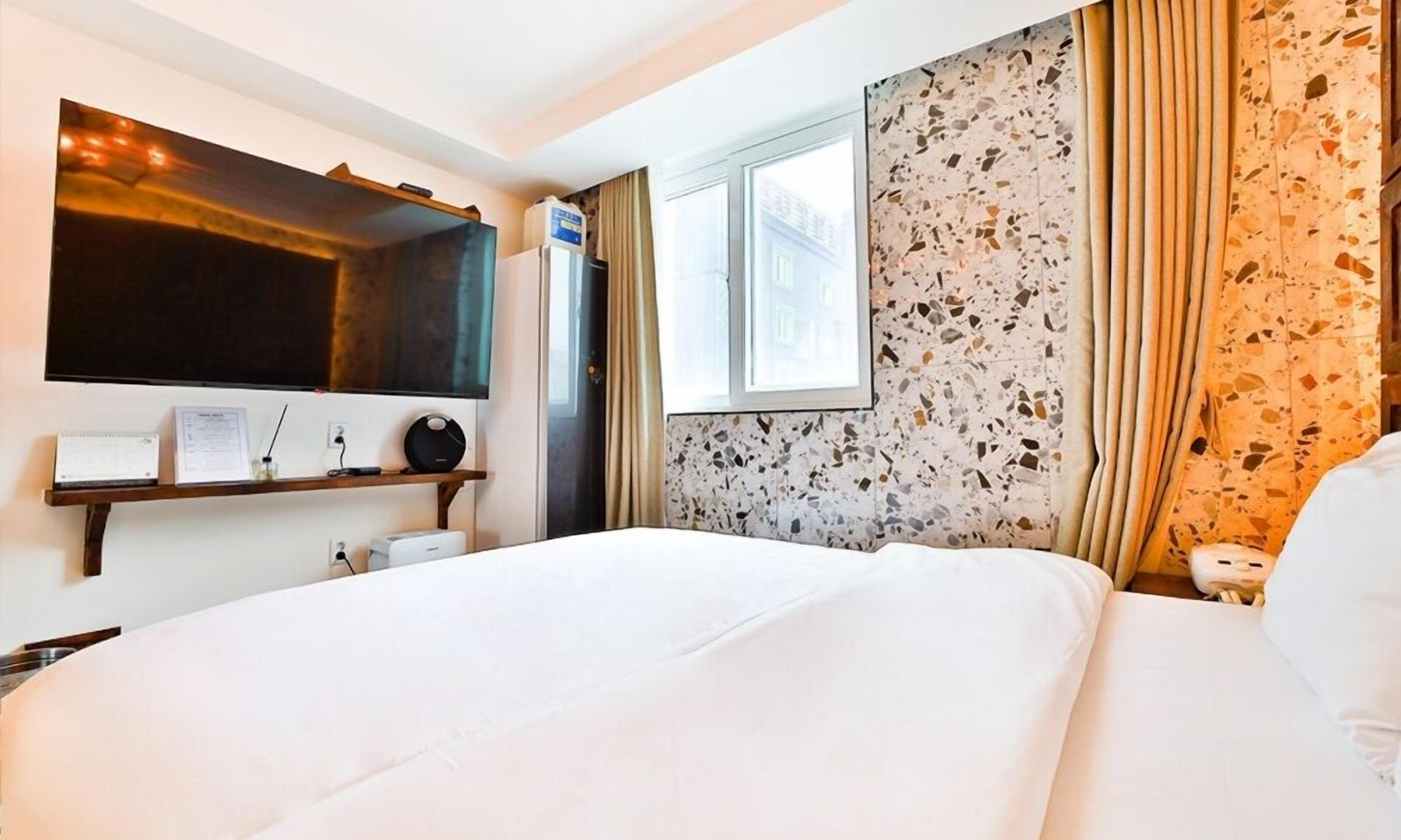 Camera d'albergo con un letto, una TV a parete, un piccolo ripiano con oggetti, una finestra e una parete rivestita in terrazzo.