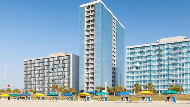 A hotel with multiple floors, a modern design, and a beachside location with sun umbrellas and lounge chairs. - One Bedroom Condo, Myrtle Beach 3317100 (Myrtle Beach)