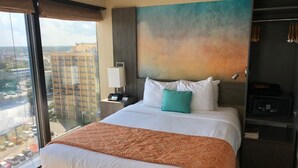 A hotel room with a large bed, a view of the city through a floor-to-ceiling window, a nightstand with a lamp, and a safe. - One Bedroom Condo, Myrtle Beach 3317100 (Myrtle Beach)