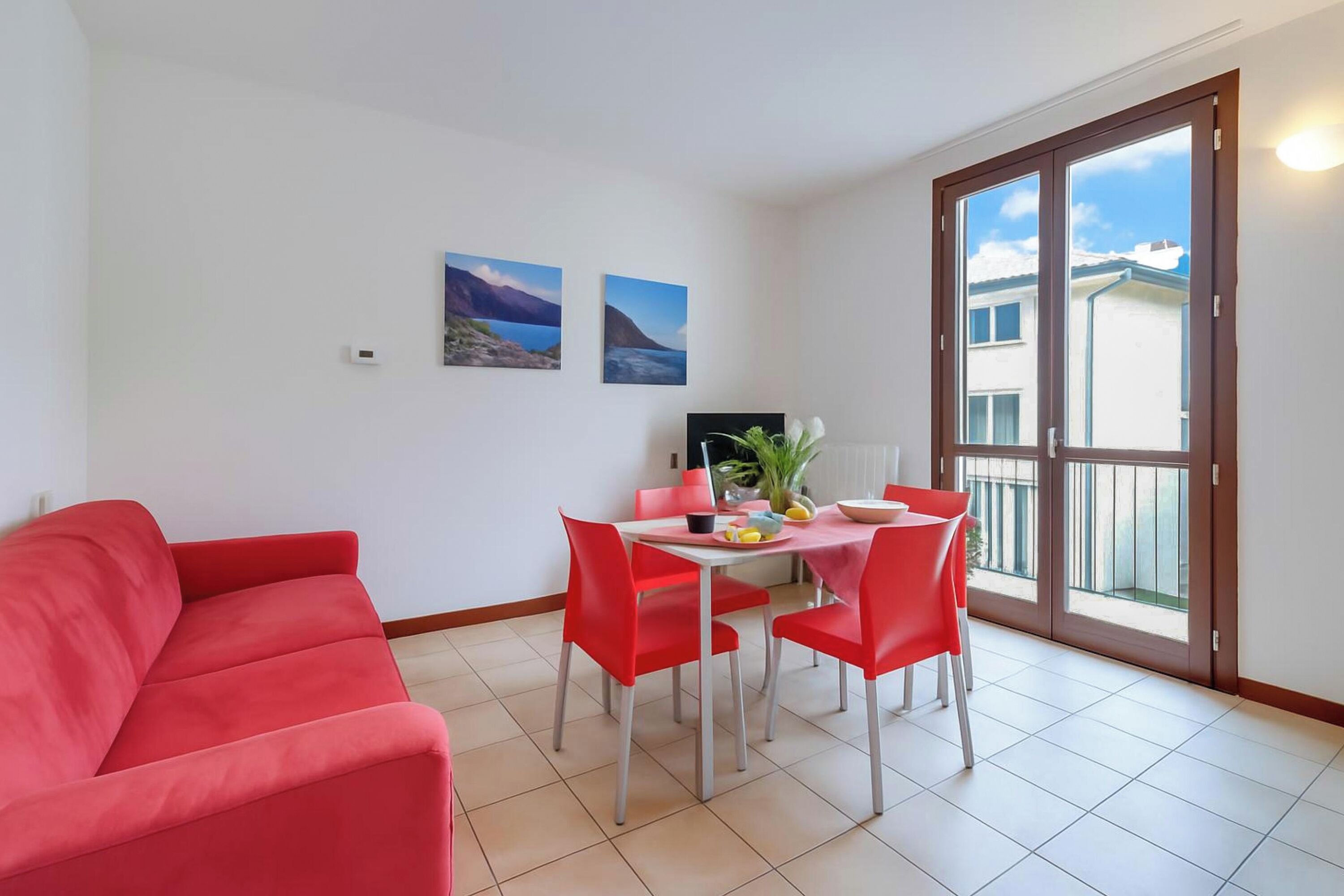 A room with a red sofa, a dining table with red chairs, two wall-mounted pictures, and a large sliding glass door to the outside.