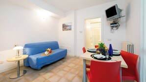 A compact living space with a blue sofa, a small dining table with red chairs, a TV mounted on the wall, and a lamp on a side table. - Porto Letizia Venezia 6, Porlezza, Italy (Porlezza)