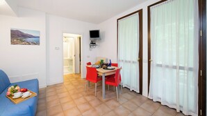 A room with a blue sofa, a dining table with red chairs, a TV mounted on the wall, and a large window with sheer curtains. - Porto Letizia Venezia 6, Porlezza, Italy (Porlezza)