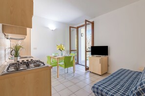 A modern kitchen with a gas stove, a dining area with a table and chairs, a TV on a stand, and a bed with a blue checkered bedspread. - Porto Letizia Mascagni 6, Porlezza, Italy (Porlezza)