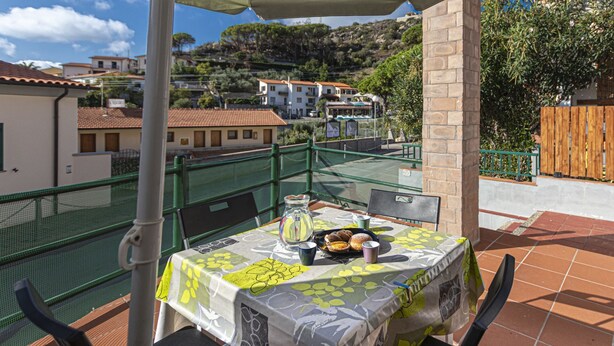A terrace with a table set for two, a green mesh fence, and a view of buildings and trees in the background. - Atena holiday apartment in Seccheto few steps away from the beach (Campo nell'Elba)