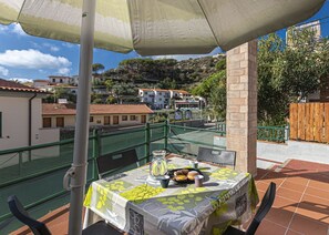 A terrace with a table set for two, a green mesh fence, and a view of buildings and trees in the background. - Atena holiday apartment in Seccheto few steps away from the beach (Campo nell'Elba)