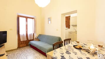 A cozy living room with a sofa, a dining table with a fish-patterned tablecloth, and a kitchen visible through an archway.