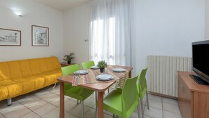 Un coin repas avec une table dressée pour quatre personnes, des chaises vertes, un canapé jaune, un téléviseur posé sur un meuble en bois et un radiateur.