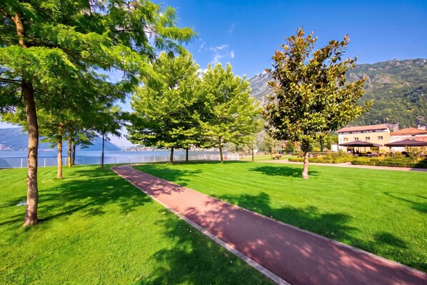 A park with a paved walkway, green grass, trees, and a view of a lake and mountains.