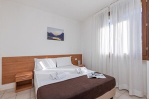 Ein Hotelzimmer mit einem Bett, einem Nachttisch, einer Lampe, einem Fenster mit Vorhängen und einem Bild an der Wand.