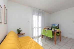Ein hellgelbes Sofa, ein Esstisch mit Stühlen, ein Topfpflanze und ein Fernseher an der Wand.