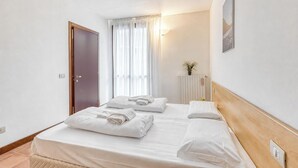 A hotel room with two beds, a wooden headboard, a door, a window with curtains, a radiator, and a wall-mounted light. - Porto Letizia Bologna 4, Porlezza, Italy (Porlezza)