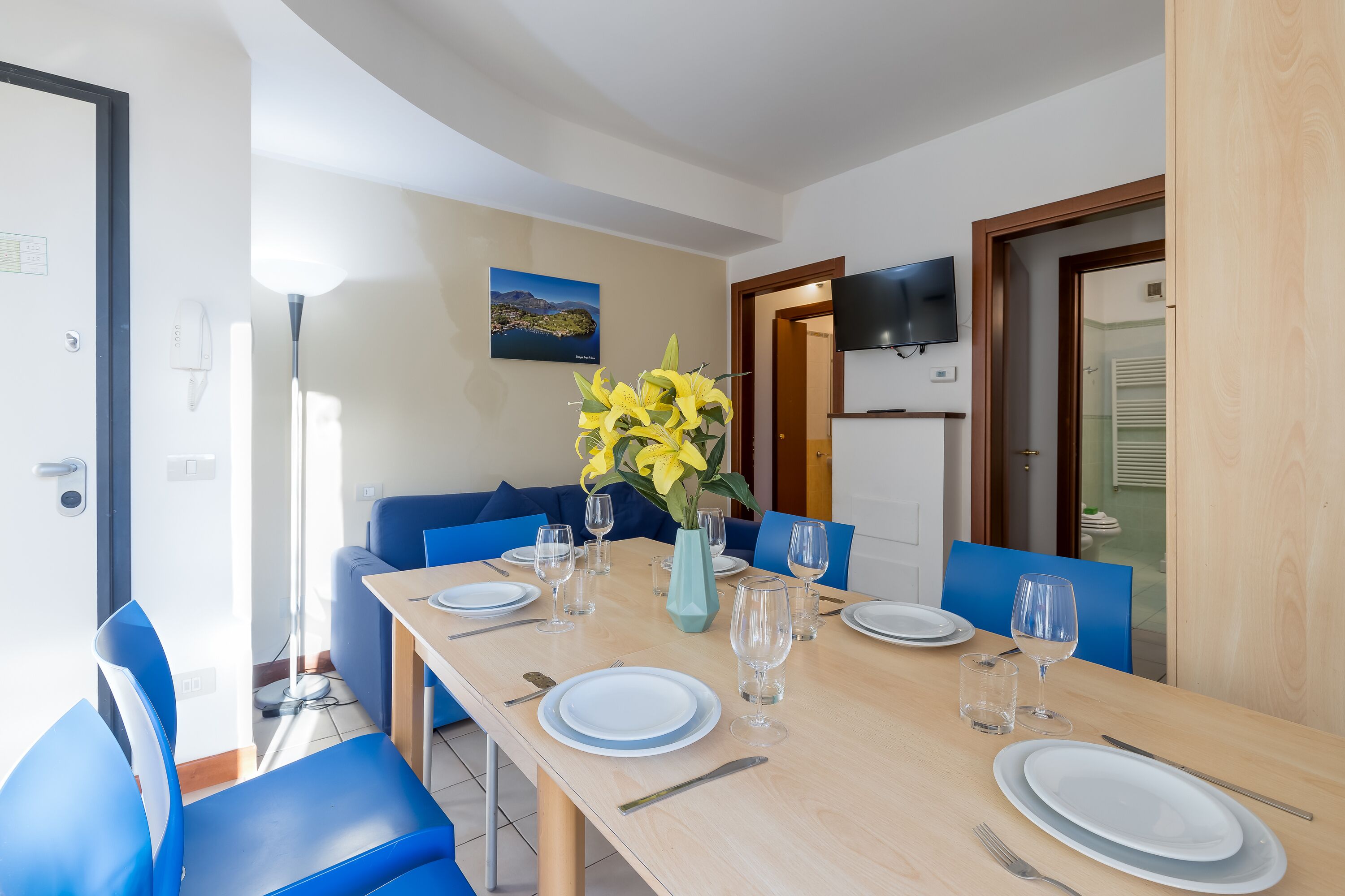 A dining area with a wooden table set for four, blue chairs, a vase with yellow flowers, and a flat-screen TV mounted on the wall.