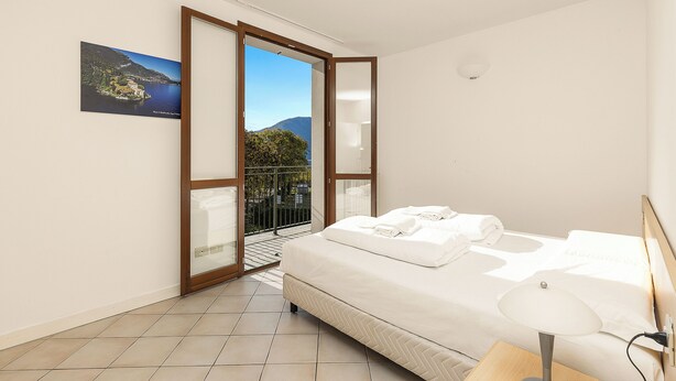 A bedroom with a bed, a bedside table, a lamp, a view of a balcony, and a painting on the wall.