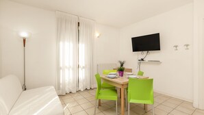 A room with a bed, a dining table with chairs, a TV mounted on the wall, and a window with curtains.