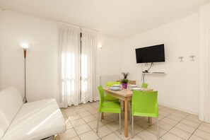 A room with a bed, a dining table with chairs, a TV mounted on the wall, and a window with curtains.