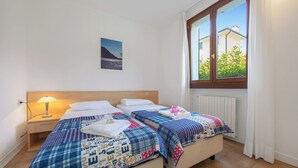 A bedroom with two beds, a nightstand, a lamp, a window with curtains, and a wall-mounted picture. - Porto Letizia Venezia 3, Porlezza, Italy (Porlezza)