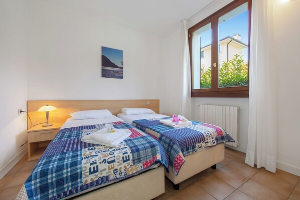 A bedroom with two beds, a nightstand, a lamp, a window with curtains, and a wall-mounted picture. - Porto Letizia Venezia 3, Porlezza, Italy (Porlezza)