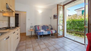 A compact kitchen with a gas stove, wooden cabinets, and a small dining table. A colorful quilted sofa is placed near a sliding glass door leading to a balcony with a view of a garden and buildings. - Porto Letizia Venezia 3, Porlezza, Italy (Porlezza)