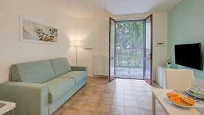 A modern living room with a sofa, a dining table with a plate of food, a flat-screen TV, and a sliding glass door leading to a garden. - Porto Letizia Mascagni 2, Porlezza, Italy (Porlezza)