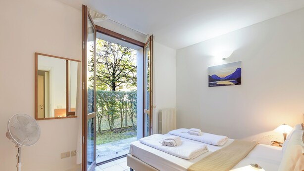 A bedroom with a bed, a bedside table, a lamp, a fan, and a painting on the wall. - Porto Letizia Leoncavallo 2, Porlezza, Italy (Porlezza)
