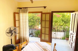 Une chambre avec un lit, une valise, un ventilateur et une vue sur une cour intérieure agrémentée de plantes et d’une clôture.