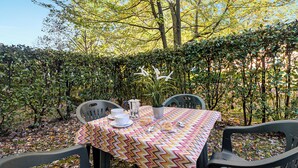 An outdoor table set with a patterned tablecloth, chairs, a coffee pot, cups, and pastries, surrounded by a hedge and trees. - Porto Letizia Beethoven 2, Porlezza, Italy (Porlezza)