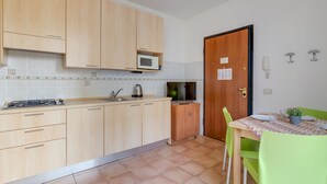 Una cocina con armarios de madera clara, un microondas, una estufa a gas y un lavamanos. Hay una mesa de comedor con sillas y una pequeña planta en maceta.