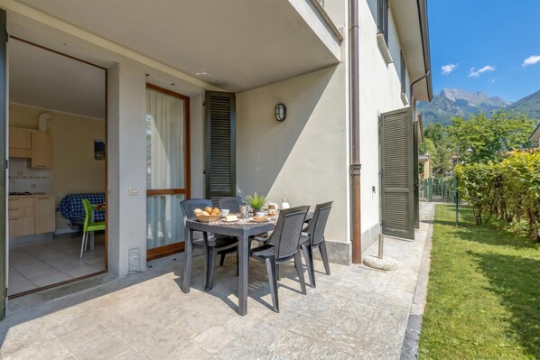 A patio with a table set for breakfast, a kitchen visible through an open door, and a mountain view in the background. - Porto Letizia Bach 3, Porlezza, Italy (Porlezza)