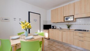 A modern kitchen with wooden cabinets, a microwave, and a dining area with green chairs and a vase of yellow flowers. - Porto Letizia Bach 1, Porlezza, Italy (Porlezza)