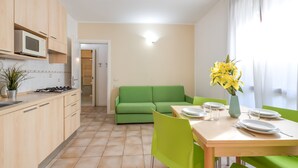 A modern kitchen with a microwave, stove, and cabinets. There is a green sofa and a dining table with chairs. A vase with yellow flowers is on the table. - Porto Letizia Bach 1, Porlezza, Italy (Porlezza)