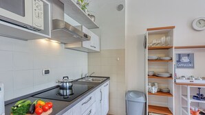 A modern kitchen with white cabinets, a black countertop, and a built-in stove. There is a trash bin and a shelf with dishes and a clock on the wall. - Porto Letizia T5, Porlezza, Italy (Porlezza)