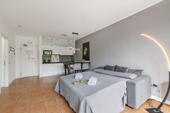 A modern hotel room with a large bed, a sofa bed, a kitchen area, and a bathroom.