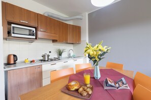 A modern kitchen with wooden cabinets, a microwave, and a sink. There is a dining table with a vase of yellow flowers and a magazine.