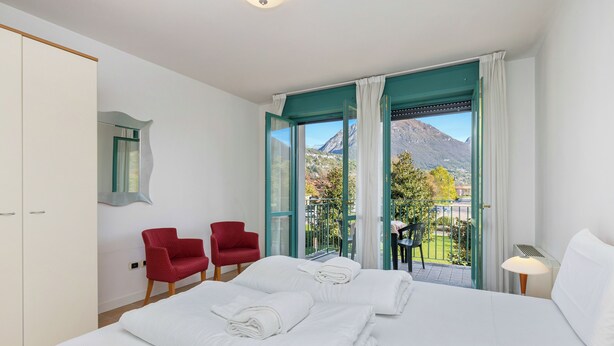A hotel room with a bed, two red chairs, a table, and a view of mountains. - Porto Letizia P17, Porlezza, Italy (Porlezza)