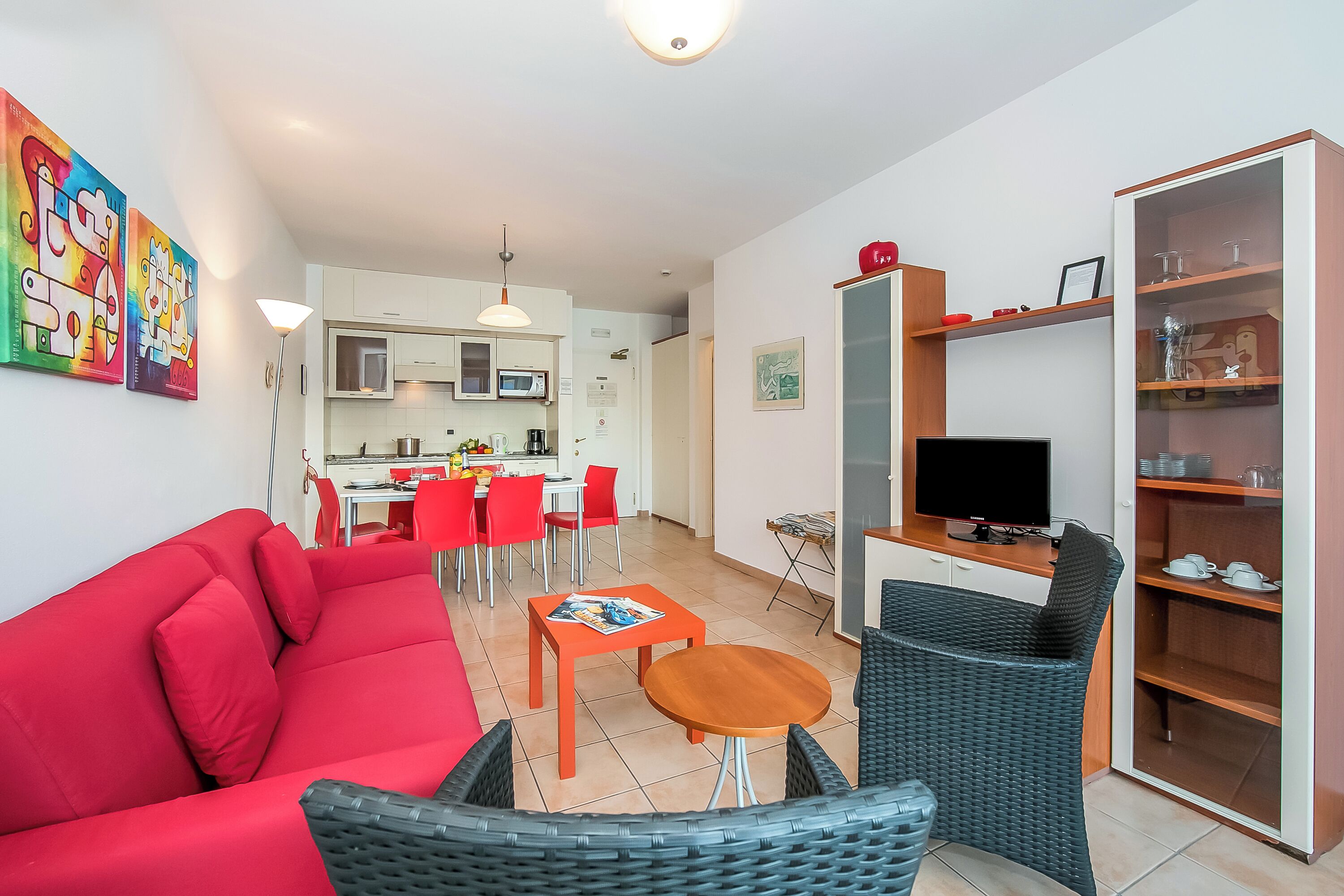 A modern living room with a red sofa, a small wooden table, and wicker chairs. A kitchen with white cabinets and red chairs is visible in the background.