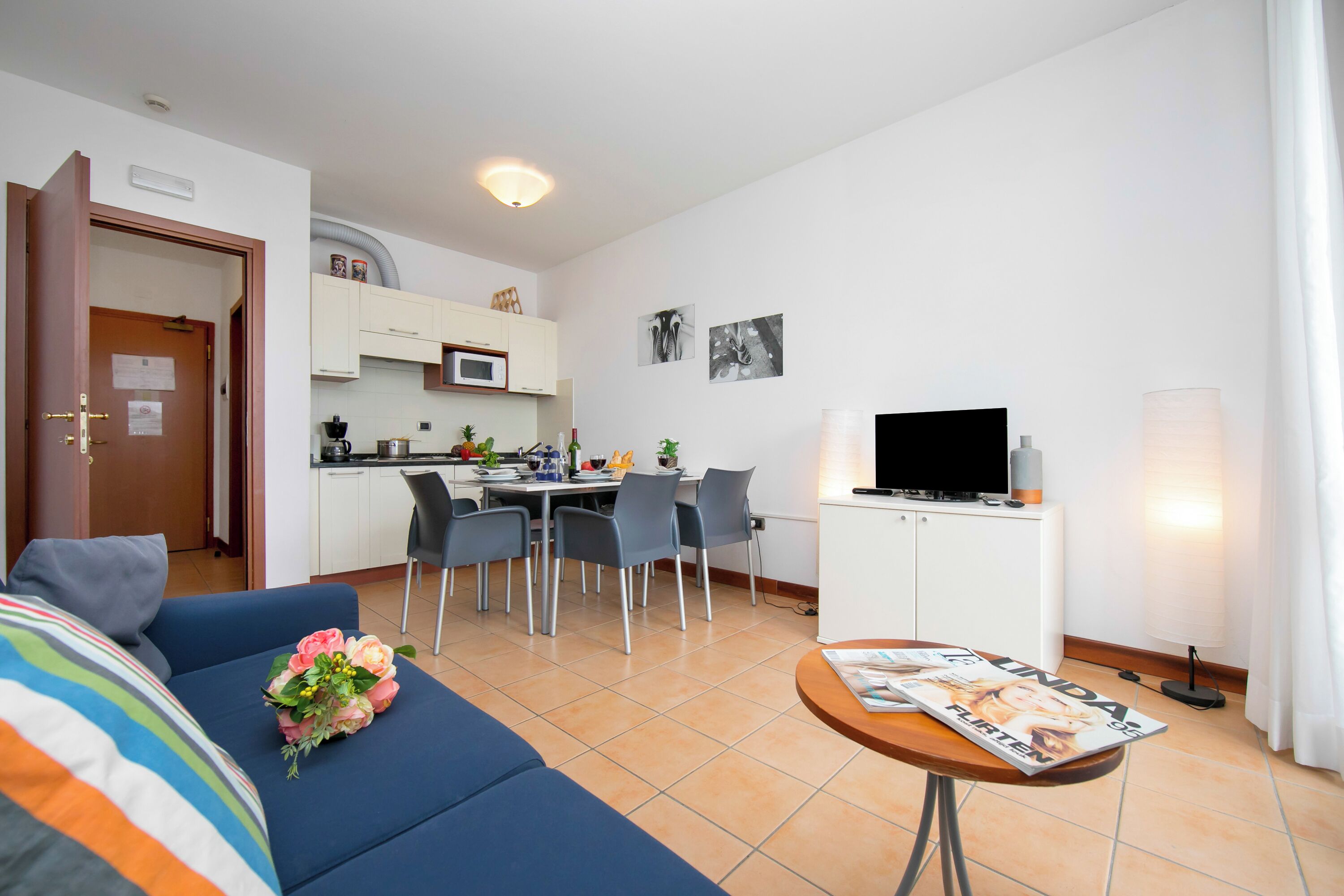 A modern living room with a sofa, dining table, and chairs. There is a kitchen area with a microwave and cabinets. A flat-screen TV is on a cabinet, and a magazine is on a small table.