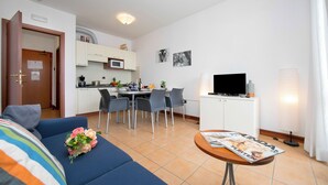 Un salon moderne avec un canapé, une table à manger et des chaises. On y trouve également un coin cuisine équipé d’un four à micro-ondes et de placards. Un téléviseur à écran plat est posé sur un meuble, et un magazine se trouve sur une petite table.