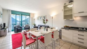 Un espace de repas moderne avec une table dressée pour quatre personnes, une cuisine dotée de meubles blancs et un balcon offrant une vue sur les montagnes.