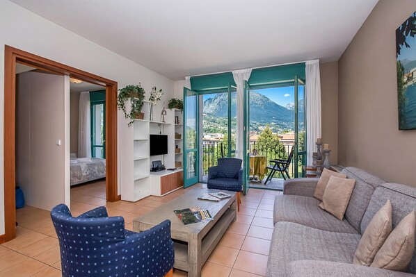 A living room with a sofa, armchair, coffee table, TV, and a balcony with a view of mountains.