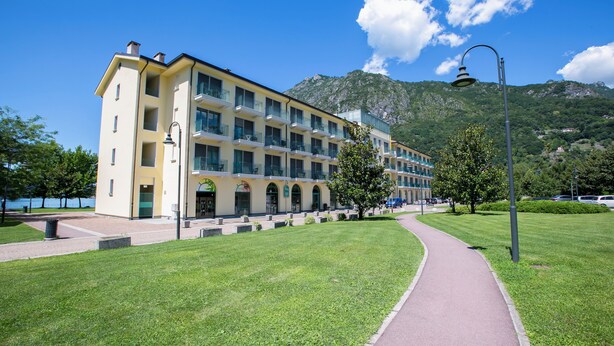 Een meerlaags hotel met een gele gevel, een verharde looppad en een bergachtige achtergrond.