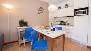 Een eetgedeelte met een tafel voor vier personen, blauwe stoelen, een vloerlamp, een magnetron en een keuken met kasten en een spoelbak.