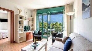 A modern living room with a sofa, armchairs, a coffee table, and a bookshelf. A balcony with a view of greenery and mountains is visible through large glass doors.