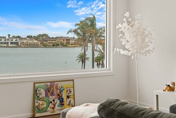 A room with a large window overlooking a body of water and palm trees, a framed Disney artwork, and a white decorative tree. - Amazing Lake View family-friendly Holiday Home (West Lakes)