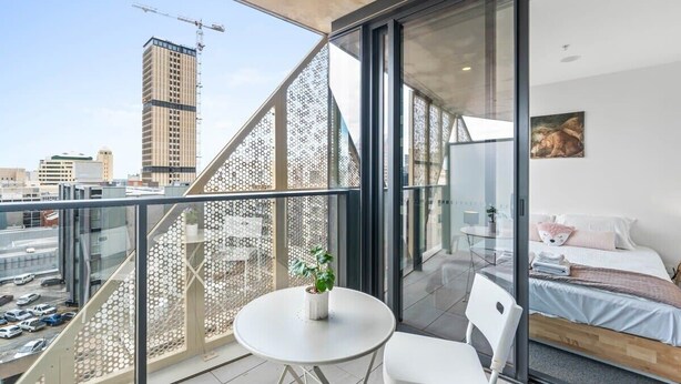 A modern bedroom with a city view, a balcony with a glass railing, and a small table with a plant.