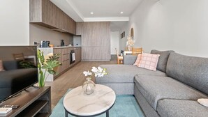 A modern living room with a grey sofa, a TV on a wooden stand, a marble coffee table, and a kitchen in the background.
