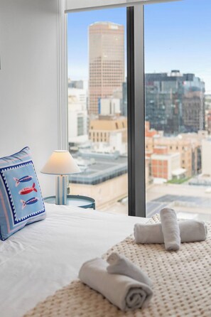 A hotel room with a bed, a pillow, a lamp, and a view of a cityscape through a large window.
