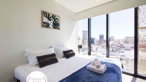 Ein modernes Hotelzimmer mit einem großen Bett, weißer Bettwäsche, einer blauen Tagesdecke und Blick auf die Stadt durch große Fenster.