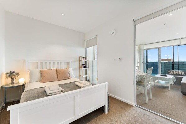 Une chambre moderne avec un lit blanc, des tables de chevet, un miroir et une vue sur un balcon aménagé avec du mobilier d’extérieur.